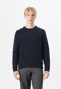 Granatowy sweter wykonany na drutach z okrągłym dekoltem, o fakturowanej tkaninie oraz ściągaczami przy mankietach i u dołu. Noszony z szarymi spodniami.