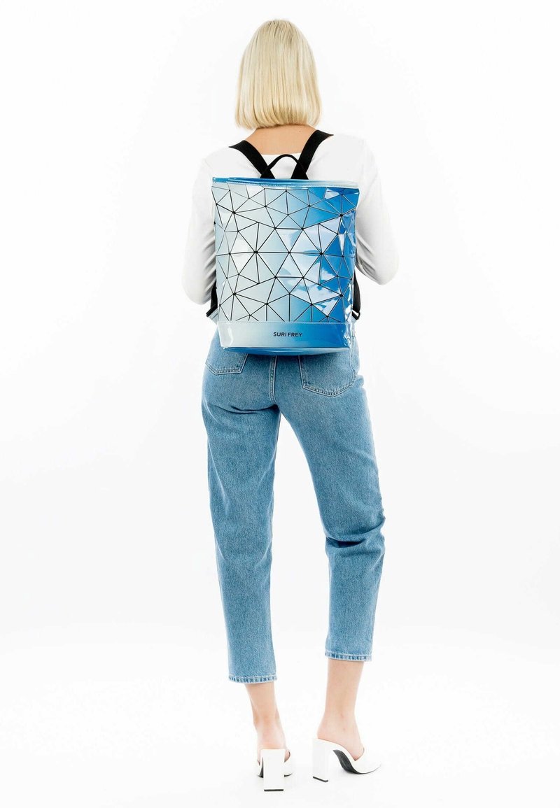 Femme aux cheveux blonds courts portant un sac à dos géométrique bleu et blanc, un haut blanc, un jean court et des chaussures blanches à talons, vue de dos.