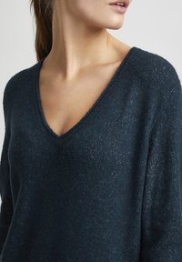 Pull en maille bleu marine avec un décolleté en V, en tissu texturé et coupe loose. Présente des bords cousus et un léger éclat.