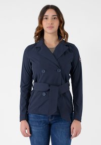 Giacca blu navy doppiopetto con cintura, realizzata in tessuto morbido. Presenta grandi bottoni e un colletto, abbinata a jeans in denim.