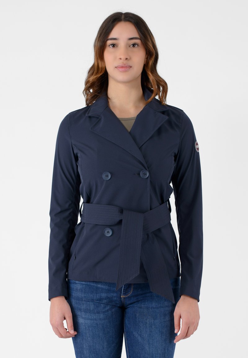 Giacca blu navy doppiopetto con cintura, realizzata in tessuto morbido. Presenta grandi bottoni e un colletto, abbinata a jeans in denim.