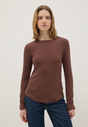 Giovane donna con capelli biondi alla lunghezza delle spalle, indossa una maglia marrone aderente a maniche lunghe e jeans blu scuro, in piedi davanti a uno sfondo chiaro e semplice.