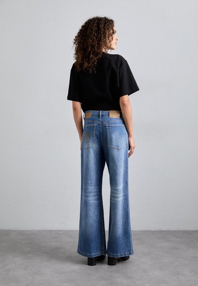 Mėlyni plačiai priglundantys džinsai su šviesiu skalbimu, turintys šiek tiek nelygumų tekstūroje ir odinę etikėtę ant juosmens. Juodas trumpomis rankovėmis viršus.