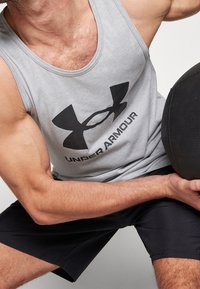 Débardeur gris sans manches avec un logo Under Armour noir, associé à un short noir. Le tissu semble léger et lisse.