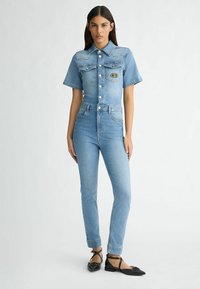 Blaue Jeansanzug mit kurzen Ärmeln, Knopfverschluss vorne, zwei Brusttaschen, hoher Taille und enganliegenden Beinen. Getragen mit schwarzen Ballerinas.