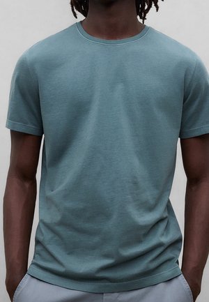 Mężczyzna w gładkim morskim t-shircie z krótkim rękawem i okrągłym dekoltem oraz jasnoszarych spodniach, stoi z rękami w kieszeniach.