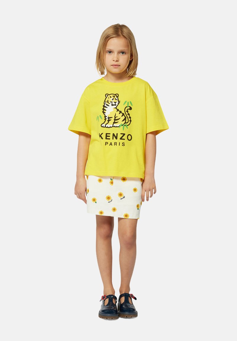 T-shirt jaune à manches courtes avec un graphique de tigre, associé à une jupe blanche ornée de motifs de tournesol. Des chaussures à brides noires complètent la tenue.