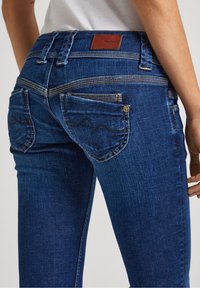 Blue denim jeans featuring a mid-rise waist, curved back pockets with zigzag stitching, and a leather logo patch on the waistband.