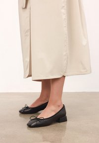 Picioare în fustă midi bej și balerini negri cu toc mic, având detalii cu fundiță, stând pe un podea din beton lustruit.