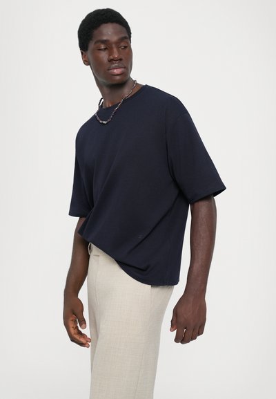 Μαύρη κοντομάνικη μπλούζα με κρουαζέ λαιμό, φτιαγμένη από βαμβάκι, σε συνδυασμό με ανοιχτό μπεζ παντελόνι. Διαθέτει άνετη εφαρμογή και λεία υφή.