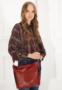 Sac à épaule en cuir rouge avec une bandoulière réglable. Texture douce et design simple. Comprend une fermeture éclair et un discret marquage à l'avant.