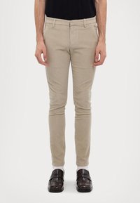 Beżowe dopasowane spodnie wykonane z teksturowanej tkaniny z płaskim przodem, ukośnymi kieszeniami i szlufkami na pasek, zestawione z ciemnymi mokasynami.