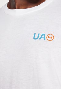 Valge puuvillane T-särk, millel on helesinine "UA" logo ja oranž ikoon stiilse "under armour" graafikaga rinnal.