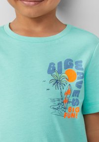 Miętowa bawełniana koszulka z graficznym wzorem przedstawiającym palmę, słońce i deskę surfingową w pomarańczowym i niebieskim kolorze.