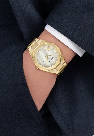 Zelta tonē Ferragamo rokas pulkstenis ar balto ciparnīcu un datumu, nēsāts uz kreisās rokas pār baltas krekla aproces un tumši zilas uzvalka piedurknes.