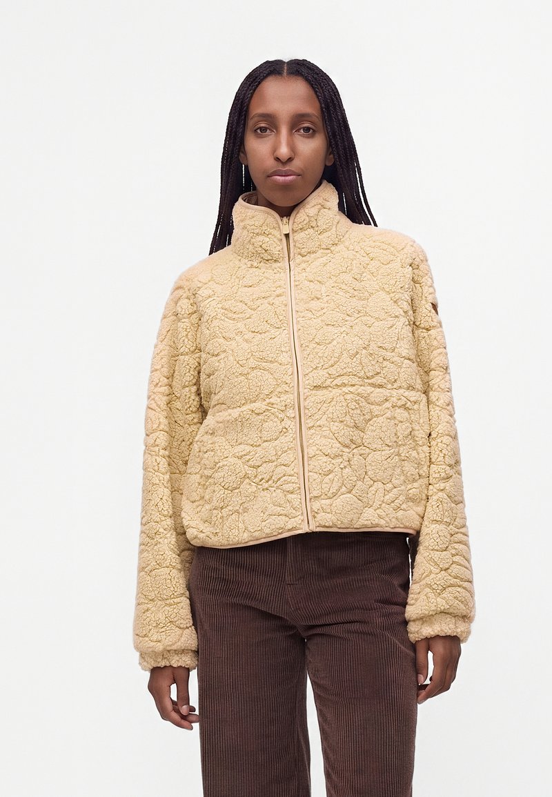 Femme aux longs cheveux tressés portant une veste polaire beige à texture fleurie et un pantalon en velours côtelé marron sur fond uni.