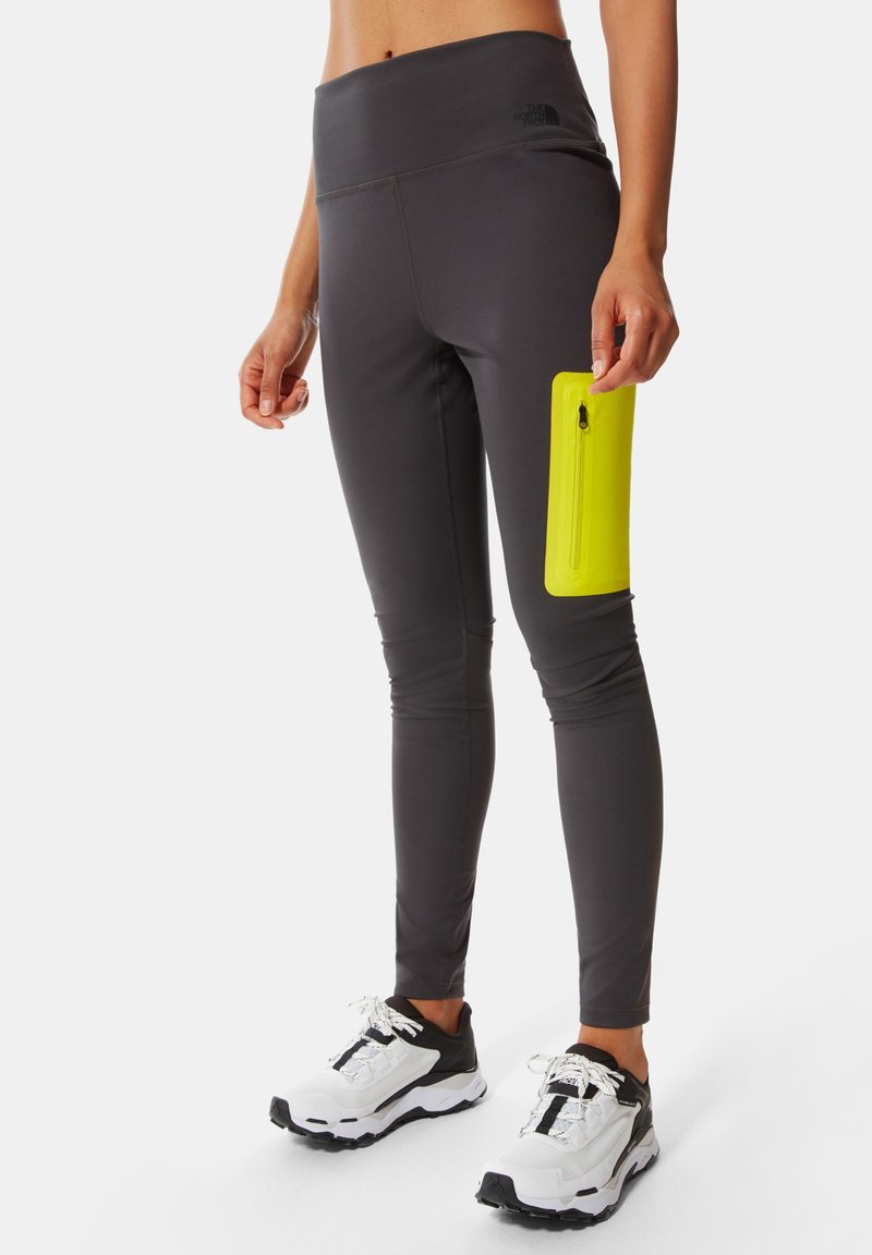 Leggings gris avec une large ceinture, dotés d'une poche zippée jaune vif sur la cuisse. Assortis à des chaussures de course noires et blanches.
