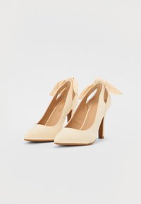 Scarpe con tacco alto beige con punta affilata, realizzate in materiale morbido, con un dettaglio a fiocco alla caviglia e un tacco sottile.