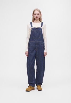 Giovane donna con lunghi capelli biondi indossa una maglia beige a maniche lunghe, una salopette di jeans blu scuro e stivali da lavoro color cuoio, in piedi davanti a uno sfondo bianco uniforme.