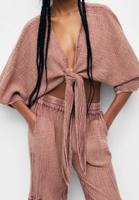 Femme portant une tenue en deux pièces texturée mauve avec un haut à col en V profond et noué devant, et un pantalon taille haute à taille élastique, les mains dans les poches.