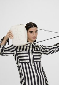 Donna con una maglietta a righe bianche e nere che tiene una borsa rettangolare bianca con la scritta in rilievo "LOLA CSDMNT" e una tracolla con il marchio.