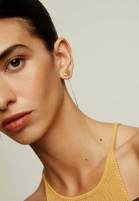 Arete de oro en forma de ola, sujeto a una cadena delgada. El sujeto lleva un top de mostaza acanalado, con piel suave y detalles sutiles.