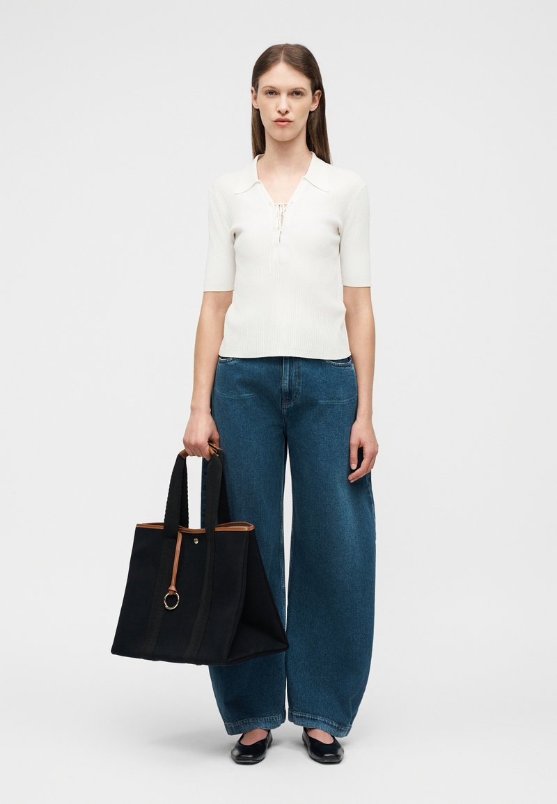 Γυναίκα που φοράει λευκό τοπ με κοντά μανίκια και φαρδιά μπλε τζιν, κρατώντας μια μεγάλη μαύρη τσάντα ώμου με καφέ περίγραμμα, στέκεται πάνω σε λευκό φόντο.