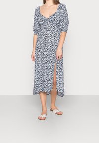 Květinové šaty v námořnické modři s bílými akcenty, krátké nafouklé rukávy, zúžený živůtek a přední rozparek. V kombinaci se světlými sandály.