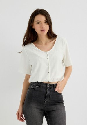 Top corto blanco con mangas cortas, tejido texturizado, cuello ancho y cinco botones en la parte delantera. Combinado con jeans oscuros de cintura alta.