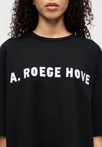 Μαύρο φαρδύ T-shirt με λευκά γράμματα σε τετράγωνο στυλ που σχηματίζουν το "A. ROEGE HOVE" στο στήθος. Λείο ύφασμα με στρογγυλεμένο ντεκολτέ.
