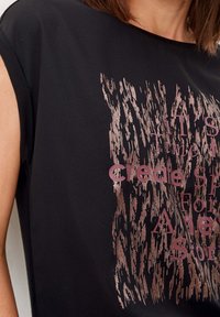 Černý top s krátkým rukávem, který má texturovaný, metalicky růžovo-zlatý grafický prvek s vytištěným textem. Měkká, hladká tkanina s volným střihem.
