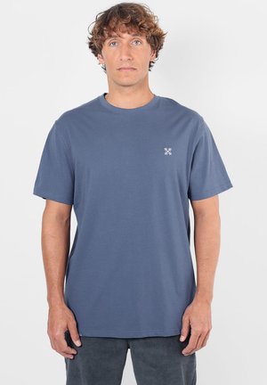 Homme aux cheveux bouclés portant un t-shirt bleu uni à manches courtes et un pantalon foncé, debout devant un fond blanc.