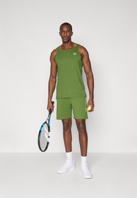 Grønn atletisk tankinis og shorts laget av lett materiale, med en rund logo og kontrastbokstaver. Hvite sneakers og tennisracket inkludert.