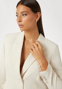 Goldkette mit klaren Edelsteinen, zarte Ring mit ähnlichen Steinen und ein cremefarbener Blazer mit tiefem V-Ausschnitt und Knopfdetails.
