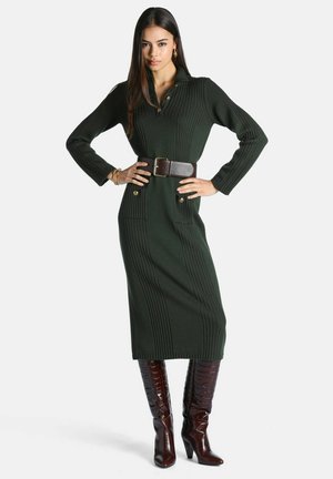 Femme portant une robe midi en maille côtelée vert foncé avec une large ceinture marron et des bottes marron montantes à talons, debout, les mains sur les hanches.
