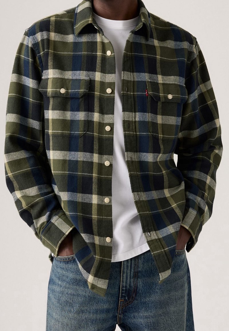 Homme portant une chemise à carreaux verte et bleue boutonnée sur un t-shirt blanc, les mains dans les poches d’un jean bleu.