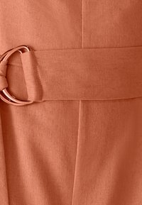 Primer plano de un cinturón de tela de color óxido texturizado con una hebilla de doble anillo de metal en oro rosado, hecha del mismo tejido.