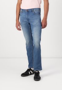 Jeans de mezclilla azul claro con un corte recto, que cuentan con un diseño clásico de cinco bolsillos y cierre de botón. Llevados con zapatillas negras.