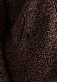 Gros plan d'une veste en polaire marron montrant un tissu texturé, une poche zippée avec bordure foncée, et un petit bouton rond noir.