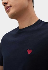 Marineblaues Baumwoll-T-Shirt mit rundem Ausschnitt und kurzen Ärmeln, das auf der linken Brustseite eine kleine rote Herzstickerei aufweist.