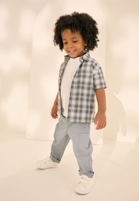 Enfant portant une chemise à carreaux à manches courtes gris et blanc, un T-shirt blanc, un pantalon cargo bleu clair et des baskets blanches.