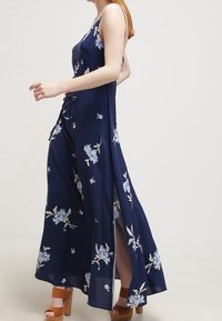 Vrouw in een marineblauwe bloemenmaxi-jurk met een zijsplit en bruine platform sandalen, die loopt tegen een effen achtergrond.