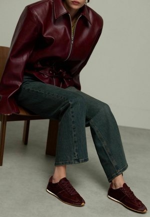 Veste en cuir bordeaux avec zip et ceinture, associée à un pantalon en denim foncé évasé et des baskets en daim bordeaux avec une semelle en gomme.