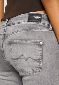 Jeans de mezclilla gris con un bolsillo trasero que presenta un patrón de costura en forma de ola distintivo y una etiqueta de cuero negra. Textura suave y diseño ajustado.