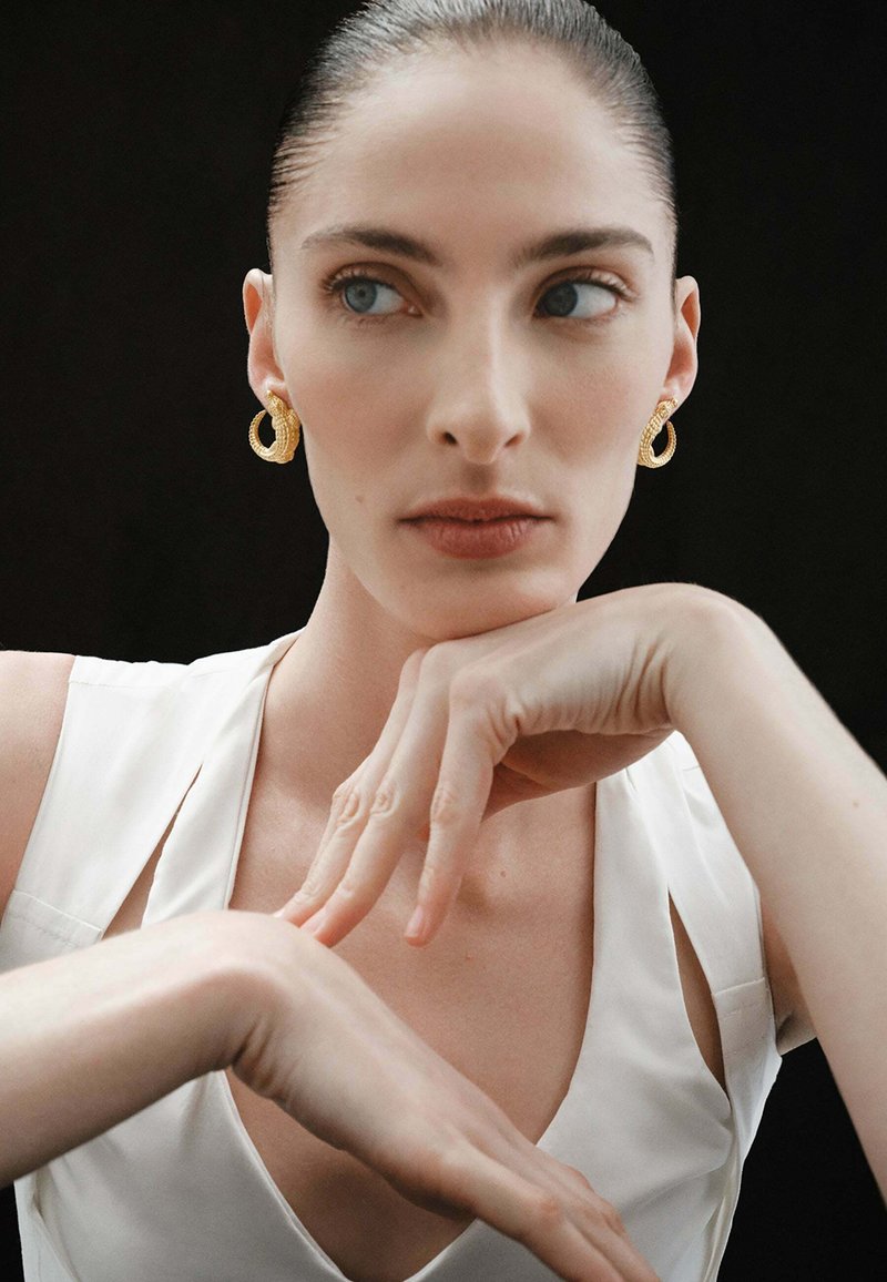 Pendientes de aro de oro con un diseño texturizado. La modelo lleva una blusa blanca sin mangas, posando frente a un fondo oscuro. Las manos están delicadamente colocadas.
