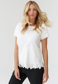 Top alb plisat cu o textură nervurată, gât rotund și hem scallopat. Asortat cu pantaloni negri, oferind un aspect curat și modern.