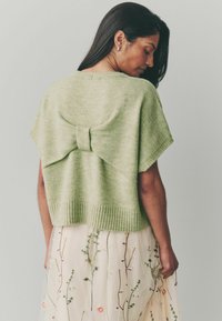Lichtgroene gebreide blouse met een brede bootnek, korte mouwen en een grote strikdetails aan de achterkant; gecombineerd met een rok met bloemenborduursel.