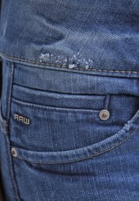 Vaqueros de mezclilla azul con un bolsillo trasero desgastado, que cuentan con un botón de metal plateado y una etiqueta "RAW". Costuras visibles y tejido texturizado.