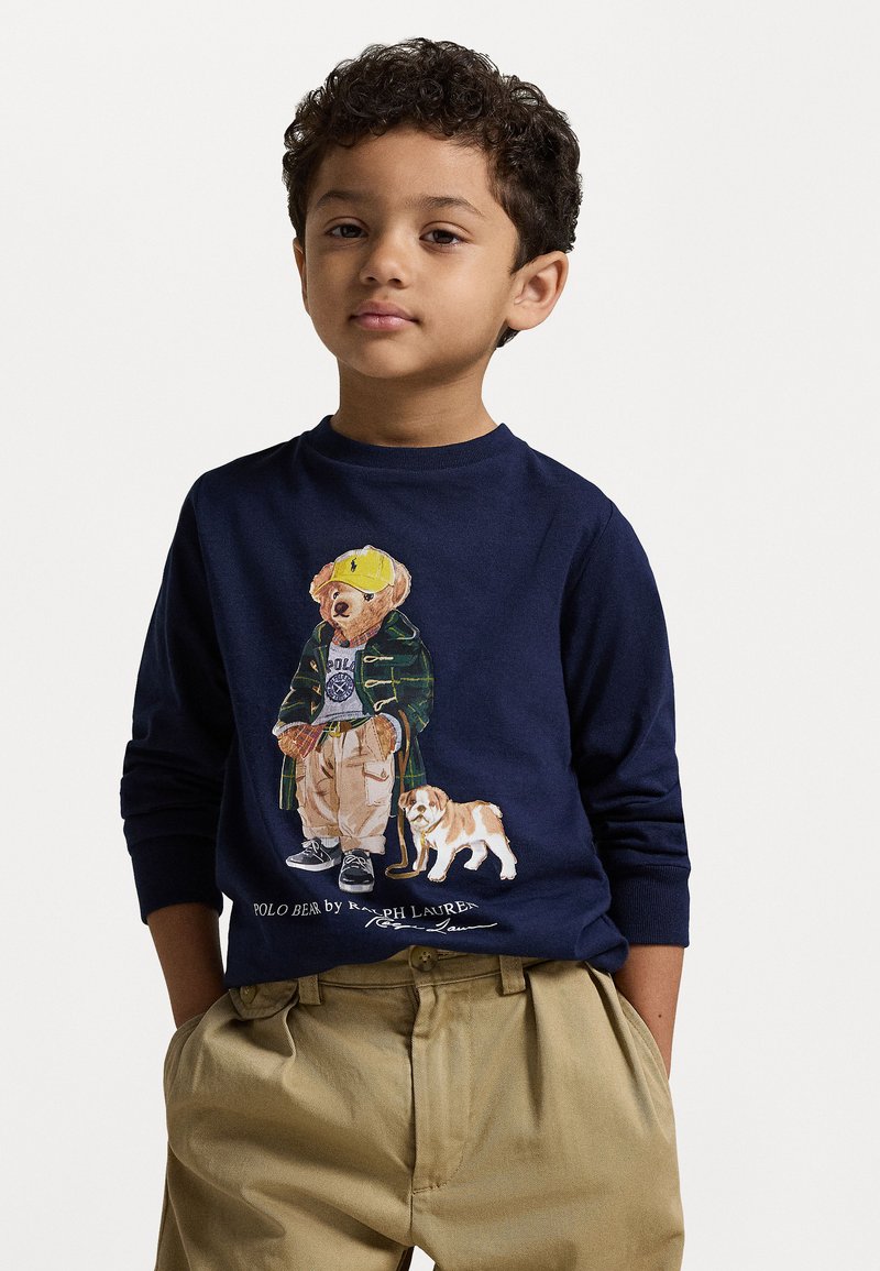 Langarm-Navy-T-Shirt mit einem gestickten Bären in einem grünen Mantel und Hund, kombiniert mit khakifarbenen Hosen. Am unteren Rand steht "POLO BEAR von Ralph Lauren."