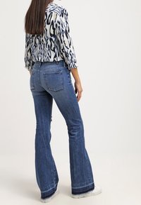 Džínové kalhoty s rozšířeným spodním lemem v středním odstínu s roztrhaným lemem a zadními kapsami. V kombinaci s vzorovanou košilí s dlouhým rukávem v modré a krémové barvě.